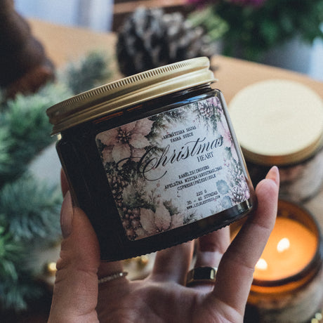 Rokā turēta CHRISTMAS HEARTH sojas vaska svece dzintara stikla traukā ar zeltainu vāku